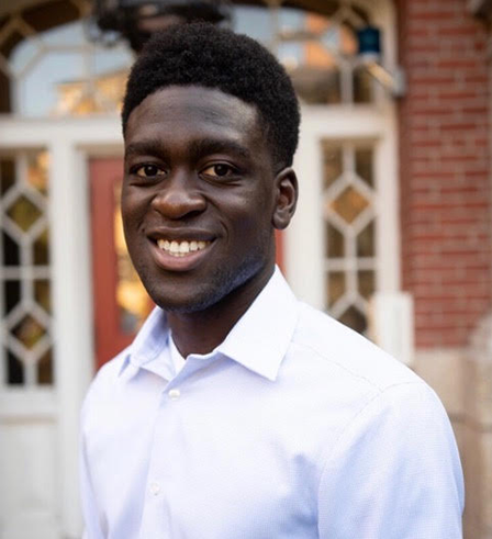 Headshot of Chris Okine in white shirt standing in front of brown building with windows