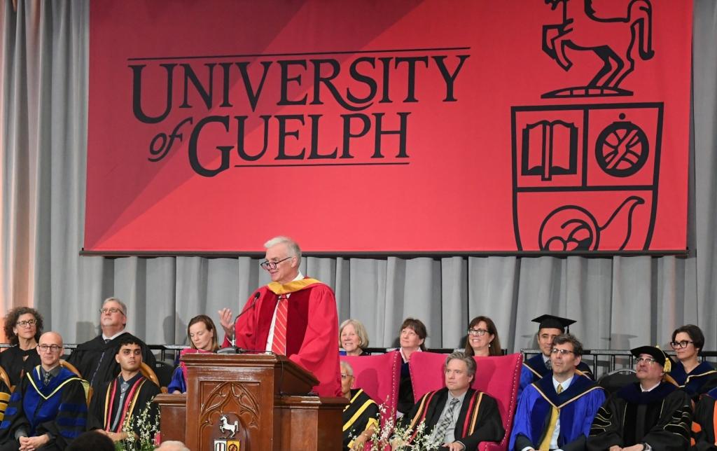 Dr. MacDougald on stage at podium with large red University of Guelph banner behind him along with people seated with graduation robes