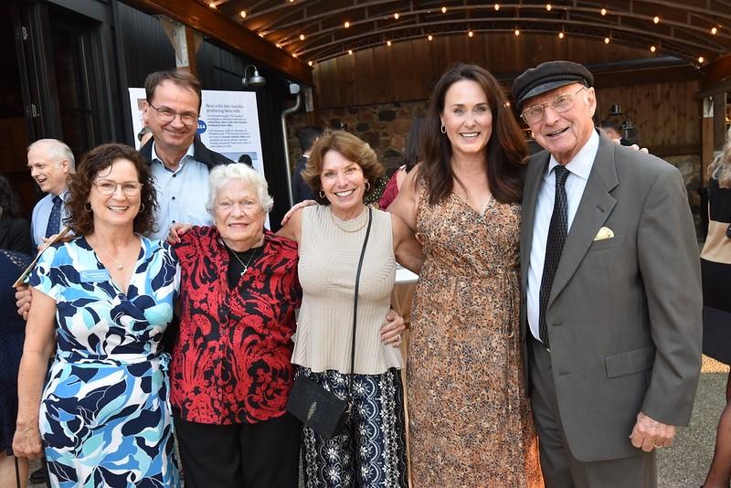 A group of six people standing together and smiling at a social event. They are posing closely in front of a warmly lit indoor setting.