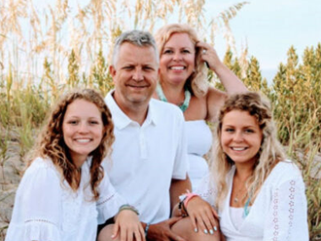 Amy Ohmer with husband and two daughters sitting outside with tall grass and trees behind them