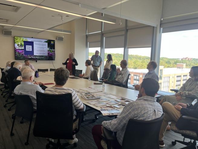 People sitting and standing around long oval table viewing a screen mounted on a wall. 