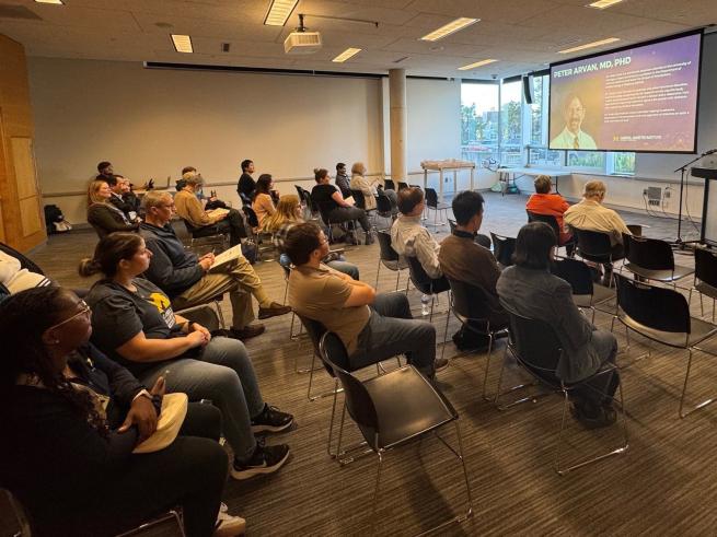 Woman stands behind podium while speaking to audience of people sitting in chairs, facing her