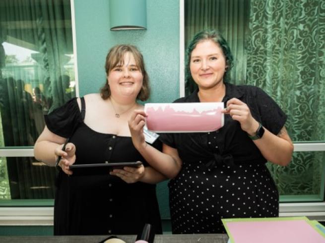Two women standing side by side, smiling with one holding up a sticker featuring a pink graph of glucose data