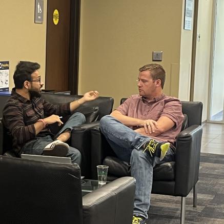 Two males sitting in chairs besides one another talking