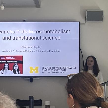 Dr. Chelsea Hepler standing behind podium with large, mounted screen to the left of her