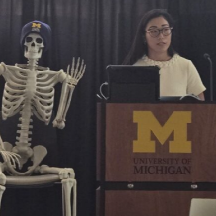 Woman with glasses stands behind a podium with the UM logo on it. A  life size skeleton with a CDI winter hat sits in a chair next to the podium, with it's hand raised in a waving position